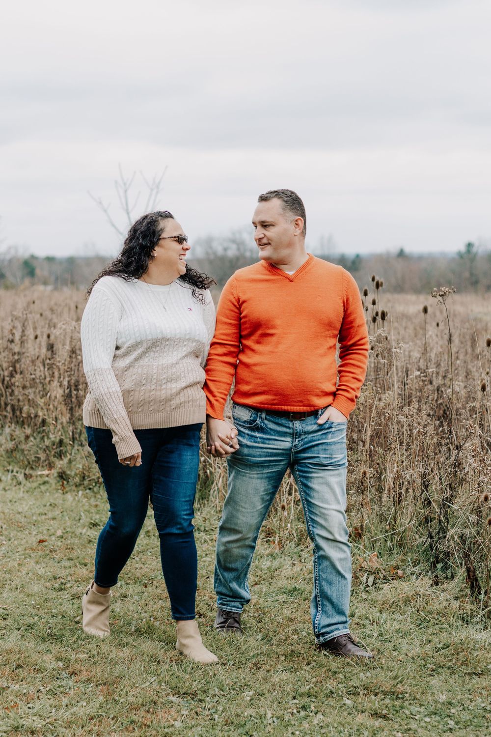 A couple in casual sweaters stands together lovingly in a field of tall dried grass on an overcast autumn day.