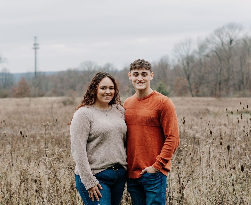 Couple in casual sweaters standing together in a winter field with a tower visible in background.