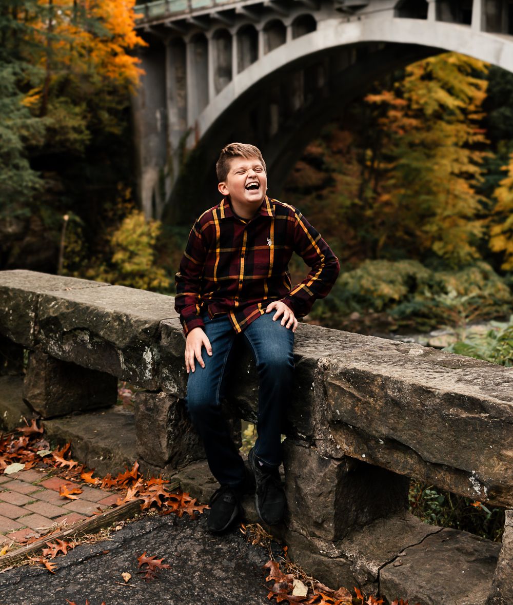 Someone in a plaid flannel shirt and jeans sits on a stone wall near a bridge during autumn with colorful fall foliage.