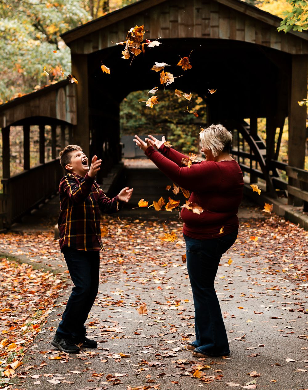 Two people tossing autumn leaves in the air while standing on a covered bridge walkway surrounded by fall foliage.