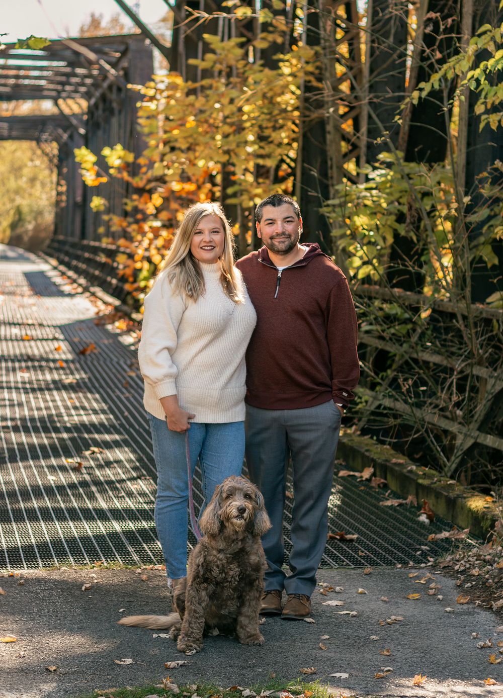 A couple walks with their brown dog on a metal walkway surrounded by autumn foliage.fall family photos northeast ohio, colleen mohr photogra
