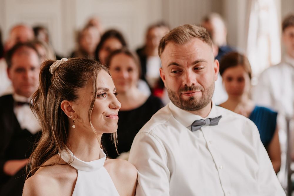 Couple smiles together during wedding ceremony with guests in background and white decorative lighting.