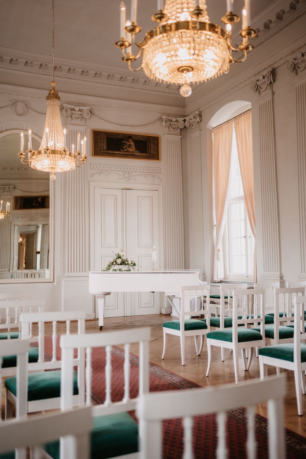 Elegant ballroom with crystal chandeliers, white paneled walls, and rows of chairs with emerald green cushions.