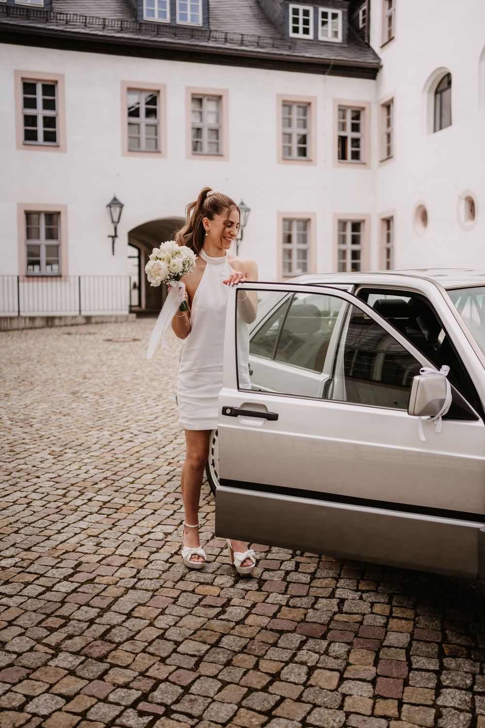 A person in a white dress stands by a vintage car on cobblestone streets outside a classic European building.