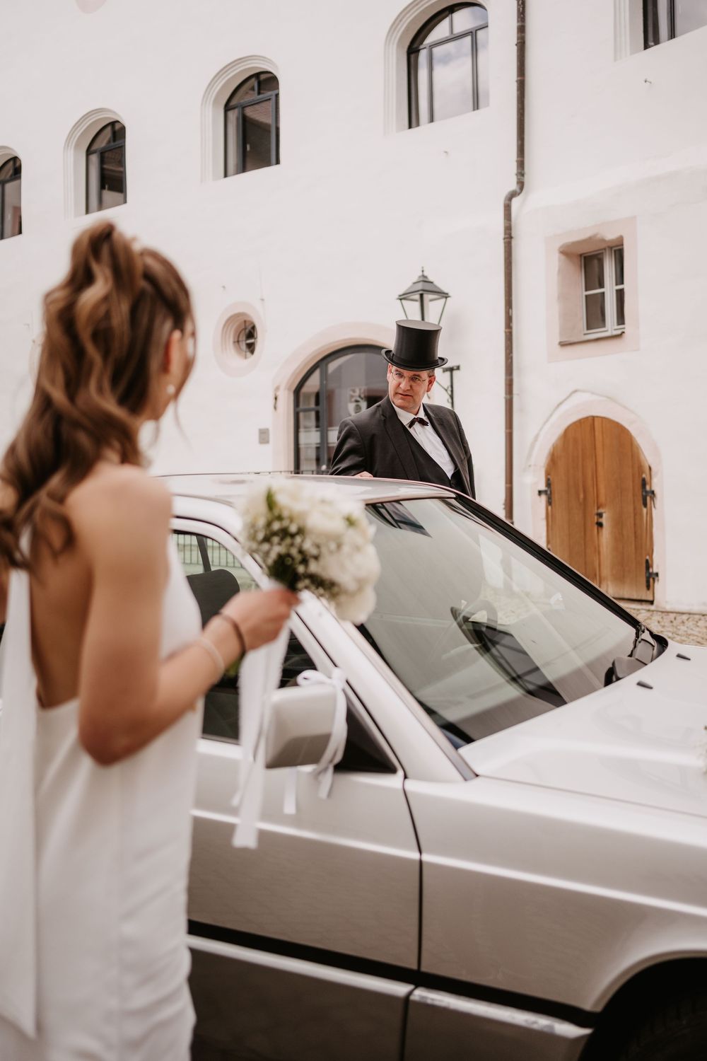 A bride in a white dress holds a bouquet while standing next to a silver car with a formally dressed chauffeur.