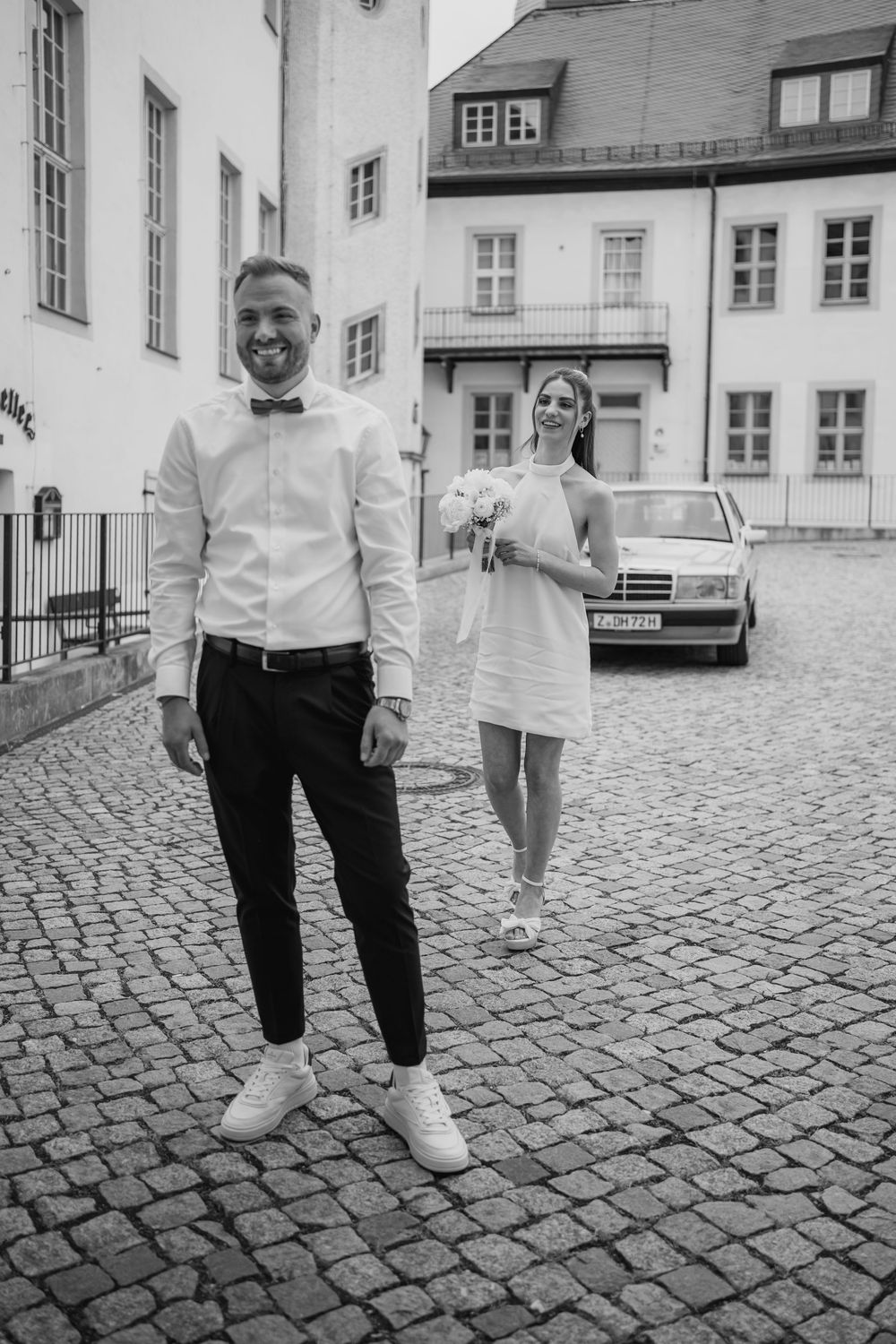 A black and white wedding photo showing a couple on a cobblestone street in front of historic European buildings.
