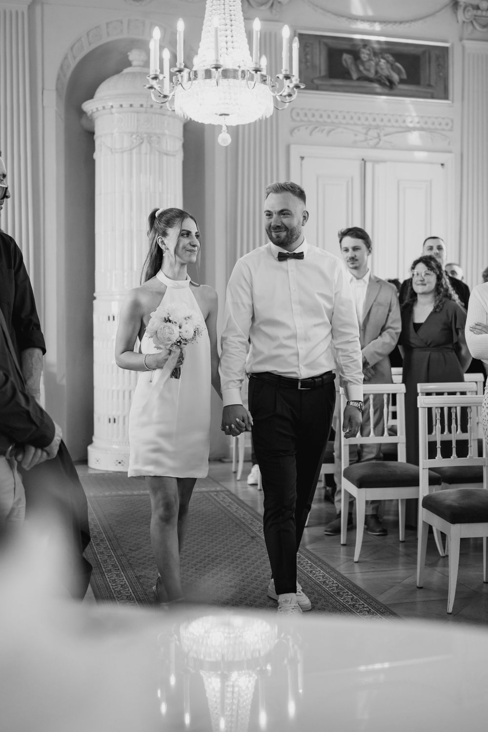 A couple in wedding attire walking together in an elegant room with white decor and a crystal chandelier.