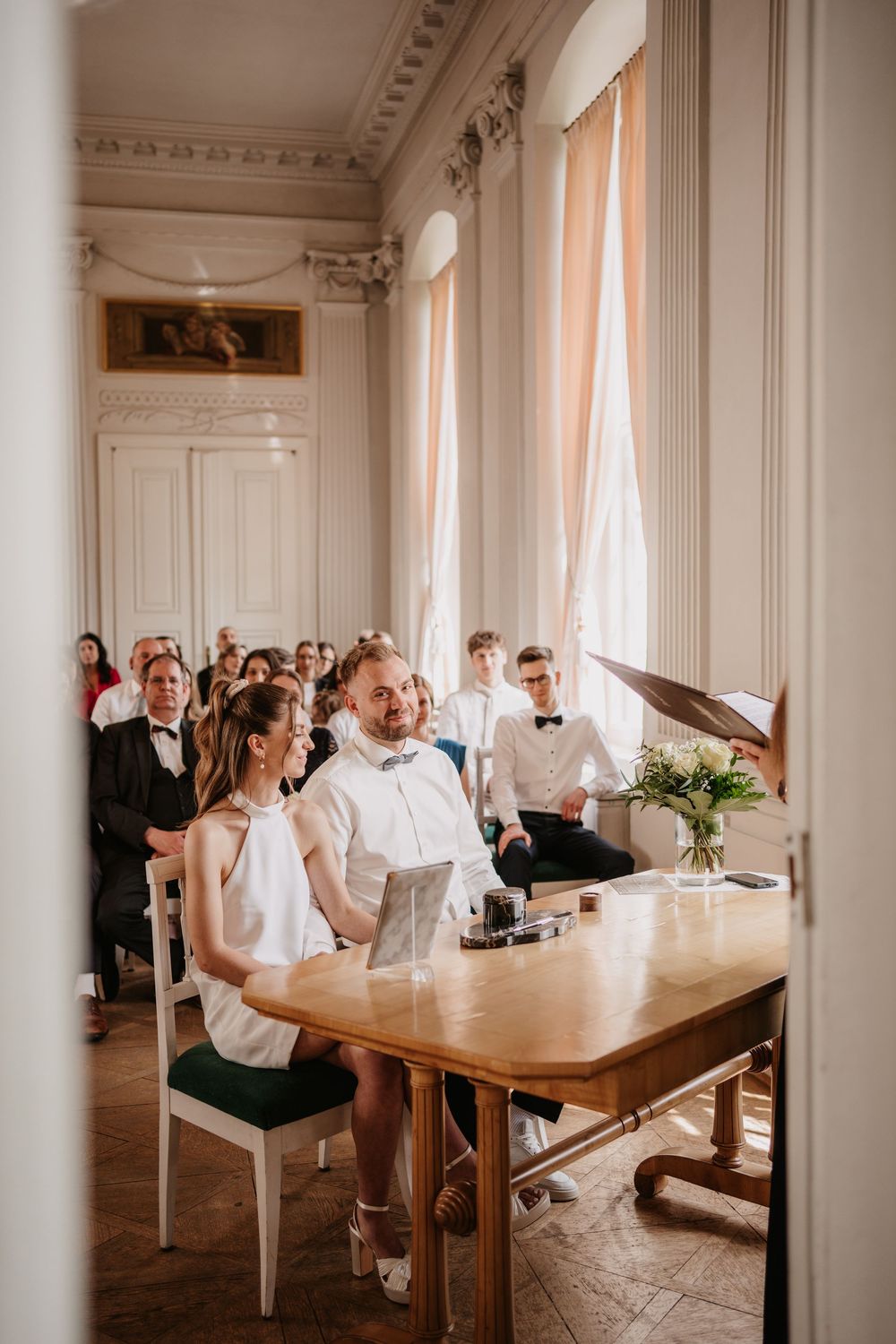 Elegant indoor ceremony setting with white decor, tall windows, and vintage wooden furniture in a bright classic room.