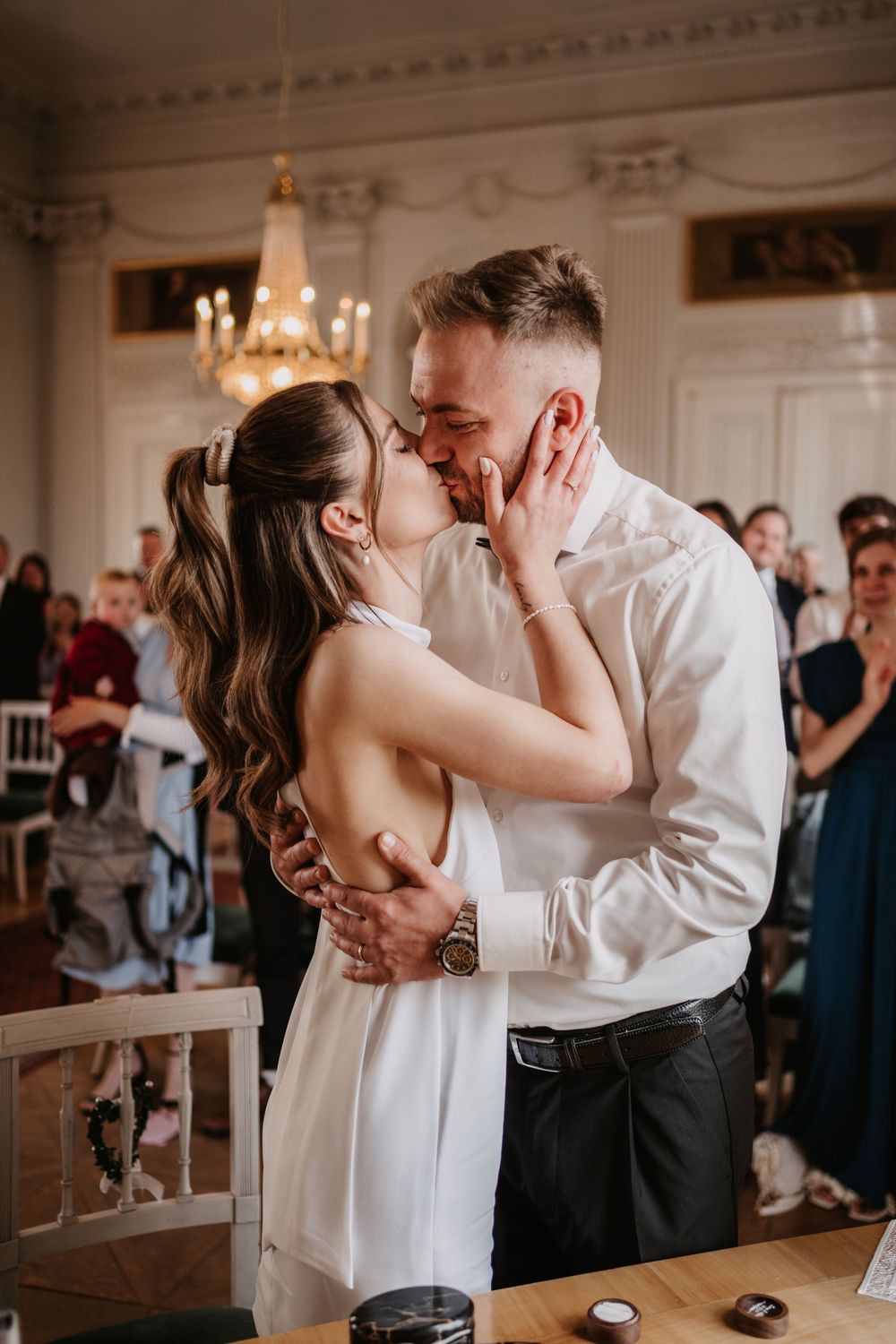 A romantic wedding moment captured in an elegant room with a crystal chandelier as the couple shares their first kiss as newlyweds.