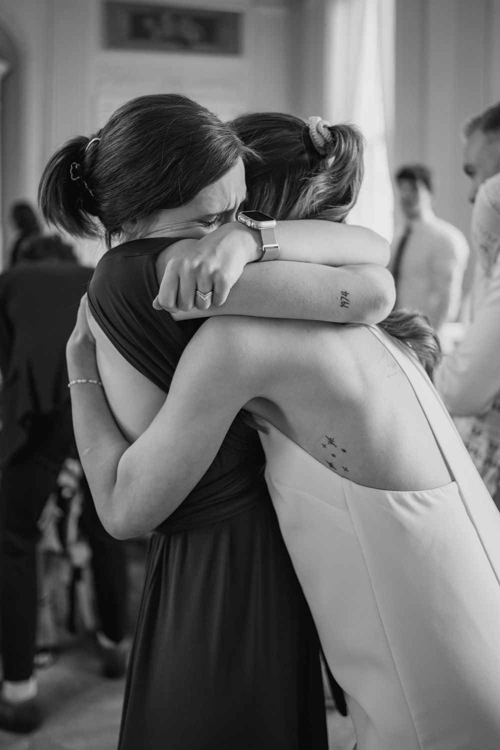 Two women share an emotional embrace at a formal event, one wearing a dark dress and one in white.