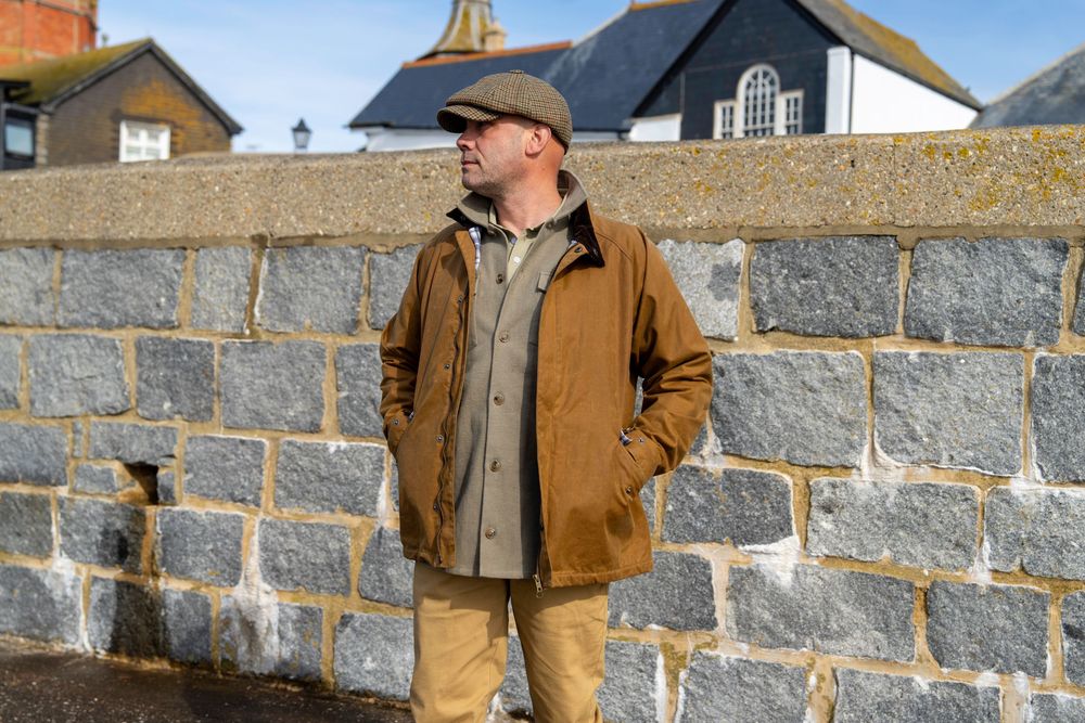 Person in brown jacket and cap standing against stone wall wearing work clothes in natural outdoor lighting.