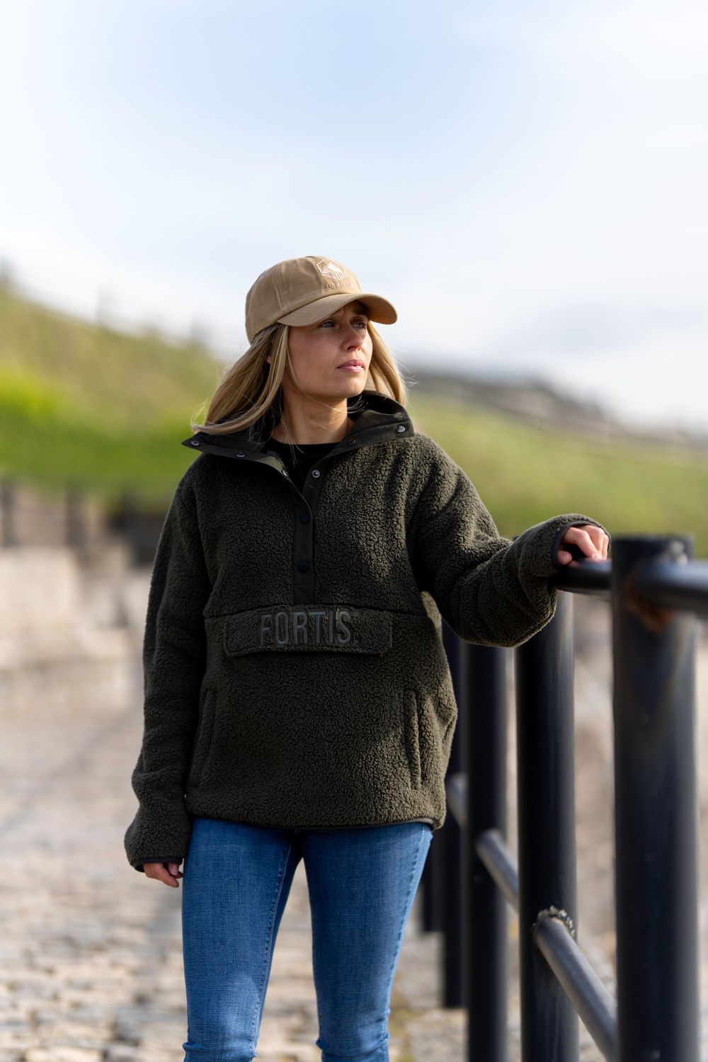 Person wearing a brown baseball cap and dark fleece pullover stands along a railing on an outdoor path.
