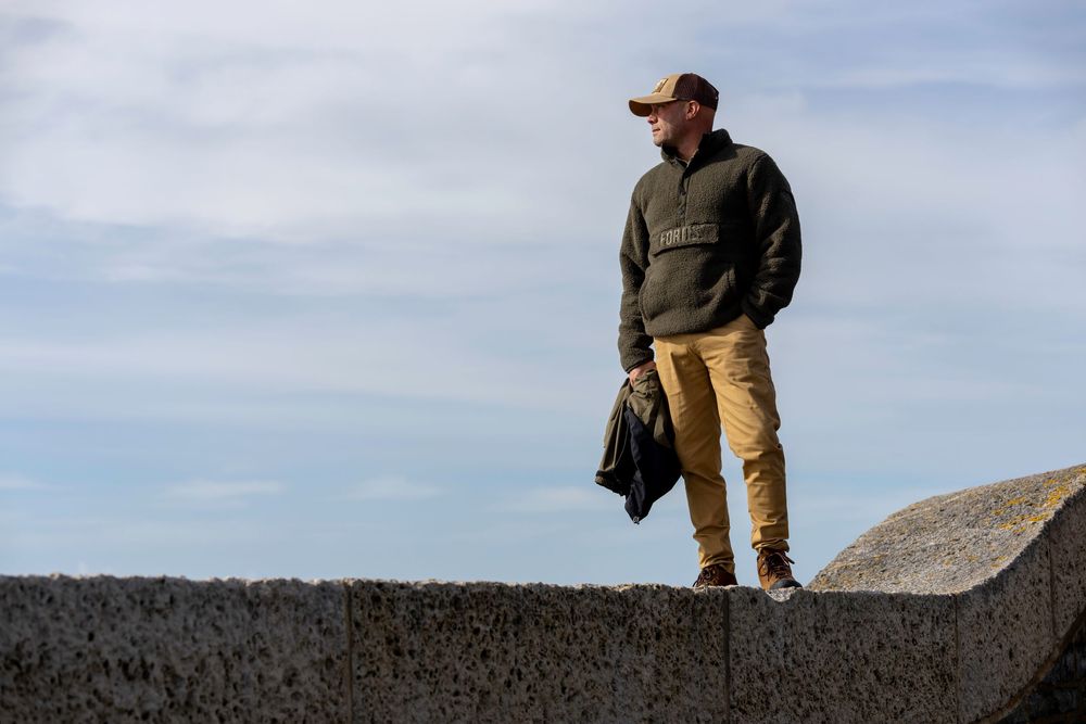 A person in casual outdoor clothing stands on a concrete ledge against a blue cloudy sky.