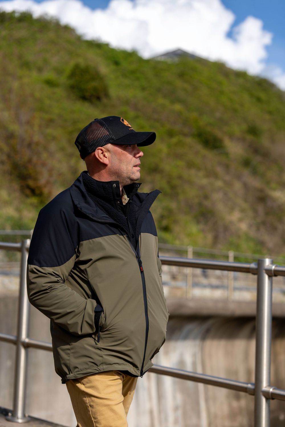 A person in a black cap and two-tone jacket stands near metal railings on a hillside path in sunny weather.