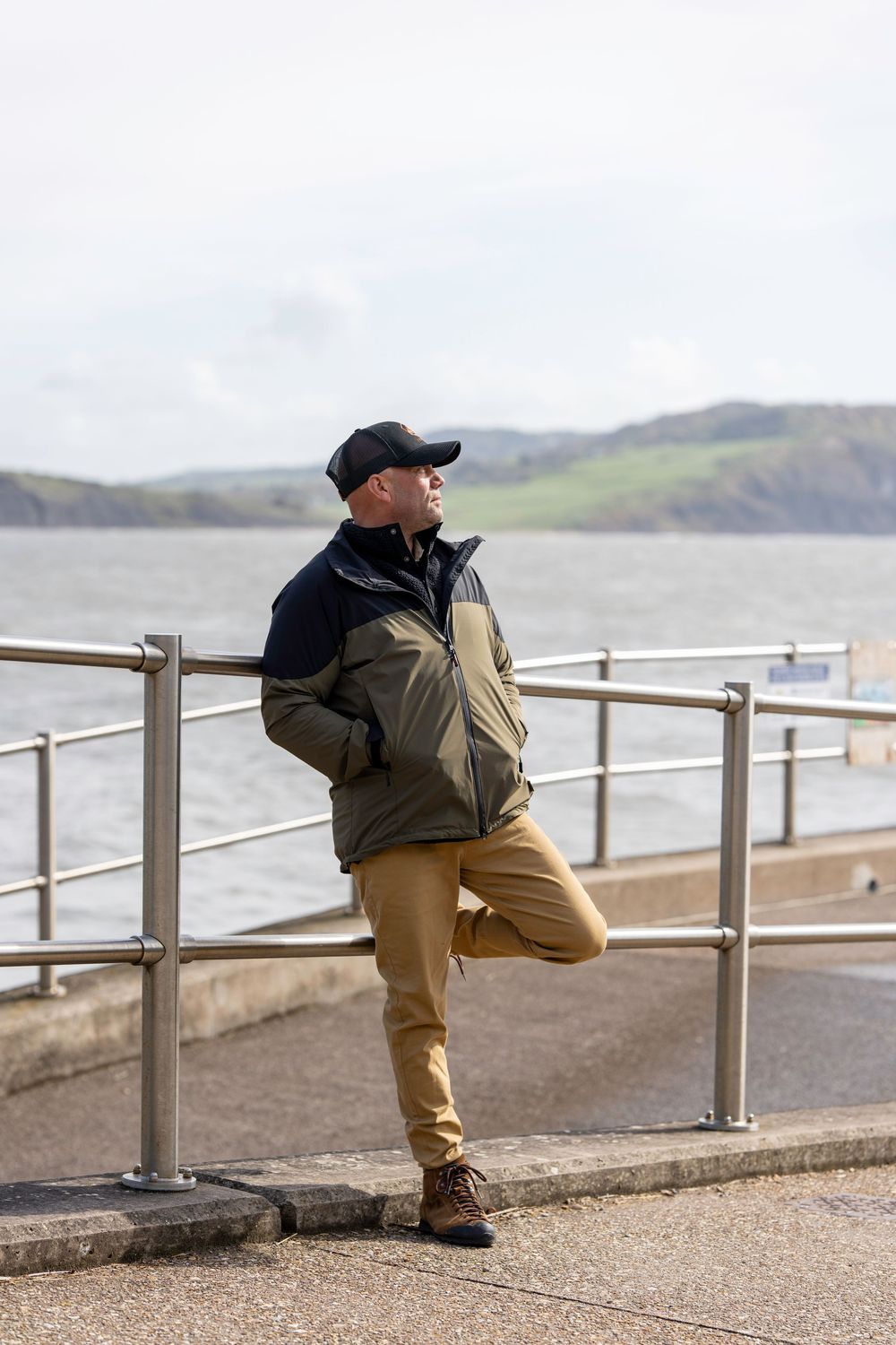 Person in khaki pants sits on a seaside railing overlooking coastal waters and distant cliffs on an overcast day.
