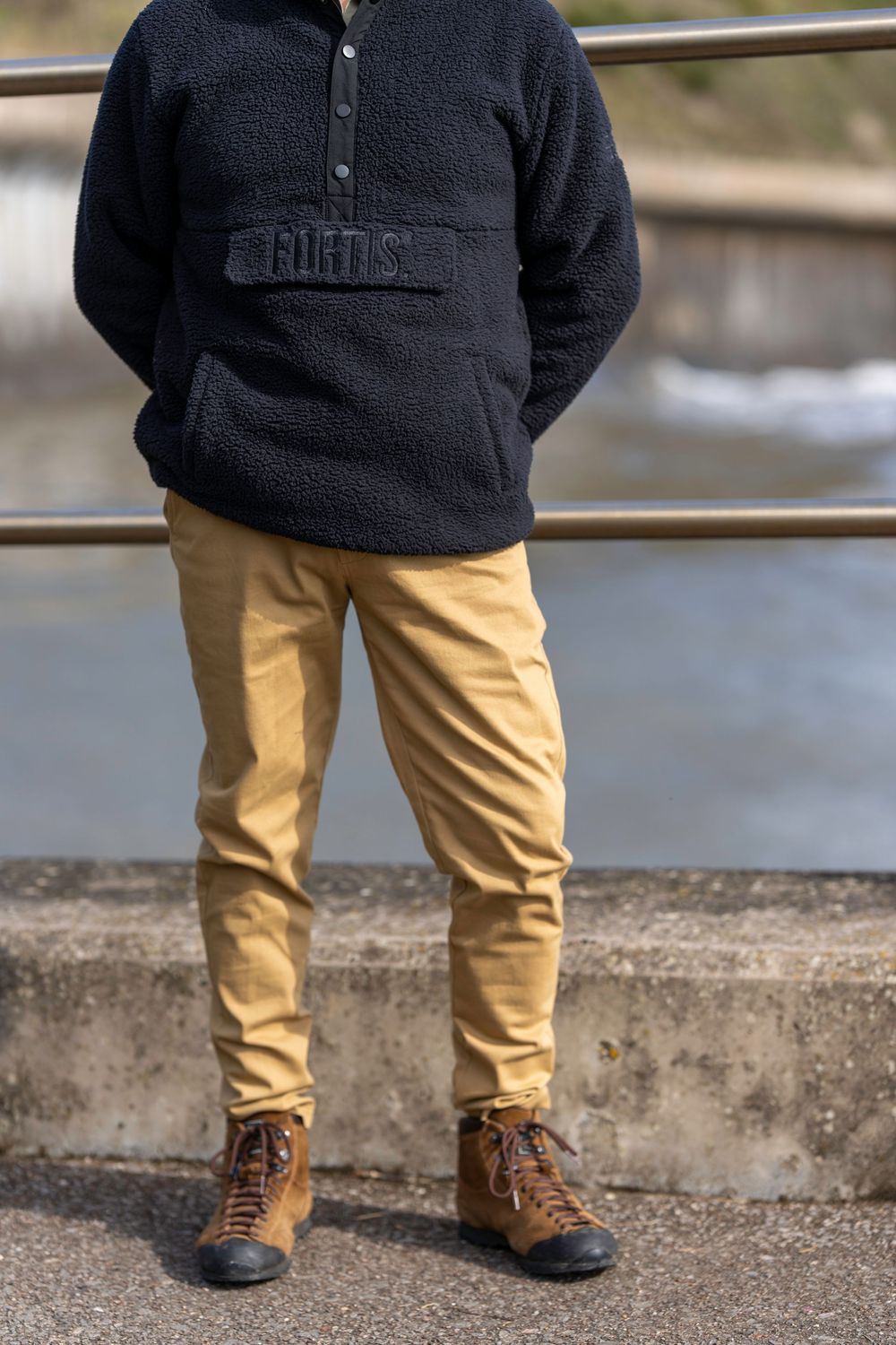 Person wearing black fleece pullover sweater with khaki chinos and brown hiking boots standing by railing.