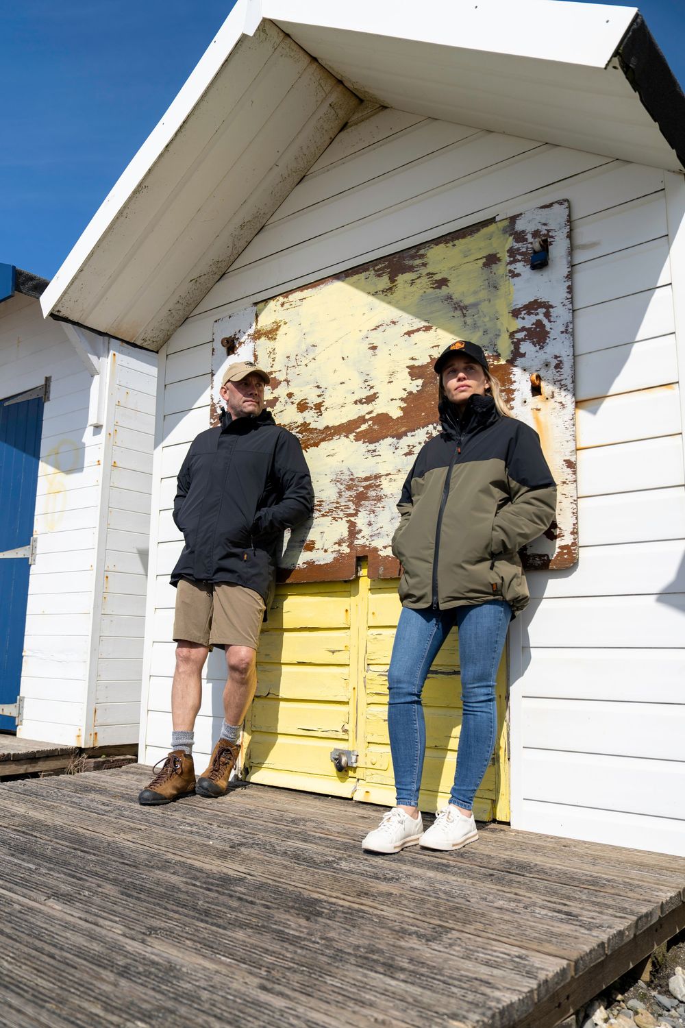 Two people in dark jackets stand on a weathered deck in front of a white building with a yellow door.