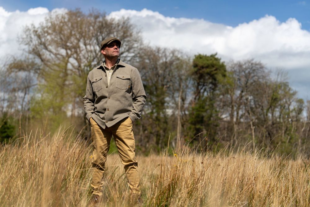 A person in khaki clothing and a cap stands among tall dried grass in a rural field on a sunny day.