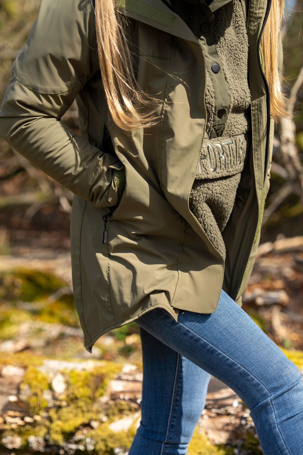 Person wearing olive green utility jacket with blue jeans standing outdoors on mossy ground in natural lighting.