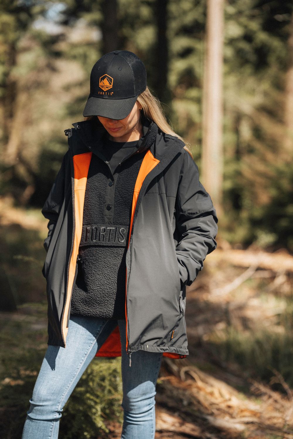 Person wearing a gray and orange jacket with light blue jeans poses in a forest setting with sunlight filtering through trees.