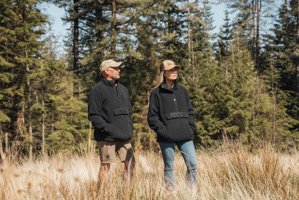 A couple walks together through an evergreen forest on a sunny winter day, surrounded by tall pine trees.