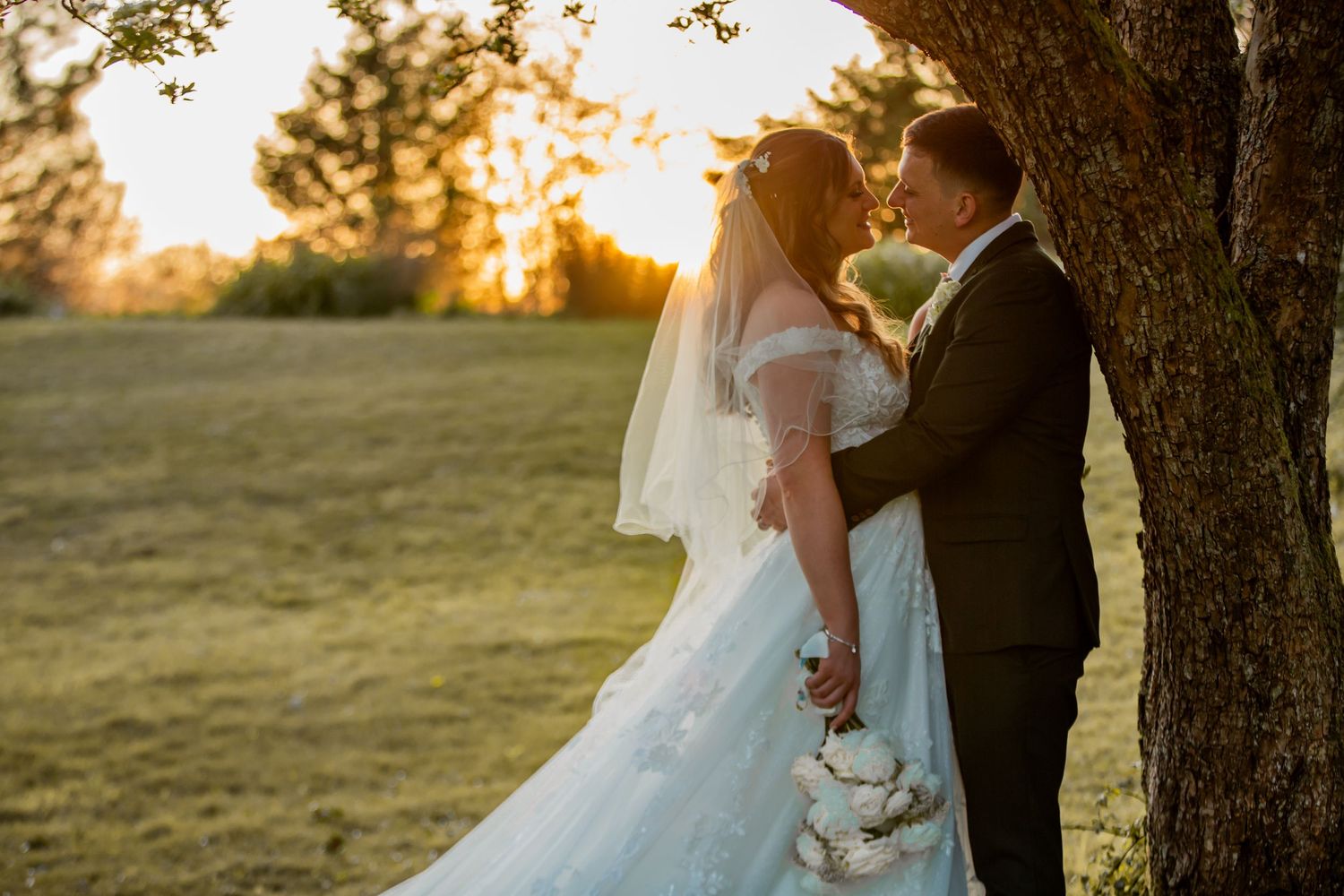 A romantic couple stands on a wooden bridge overlooking a pond with autumn trees and golden sunlight in the background.
