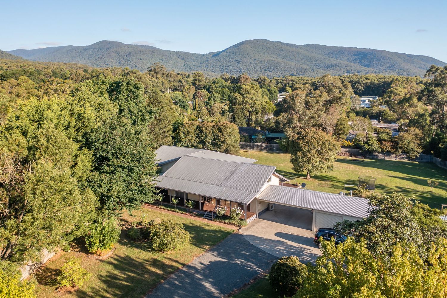 Aerial view of rural homestead surrounded by lush trees with stunning mountain ranges backdrop.
