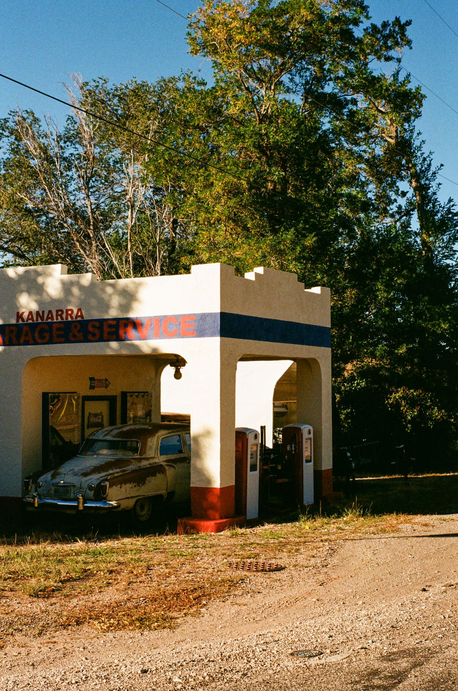 Abandoned roadside motel with white stucco buildings and trees behind it sits empty on a gravel driveway.
