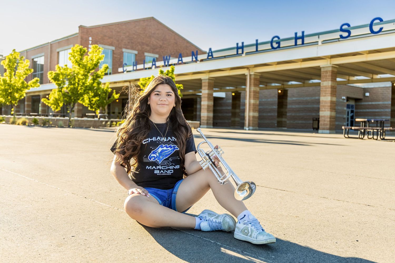 A teenage girl sits on pavement holding a trumpet in front of Cahaba High School on a sunny day.