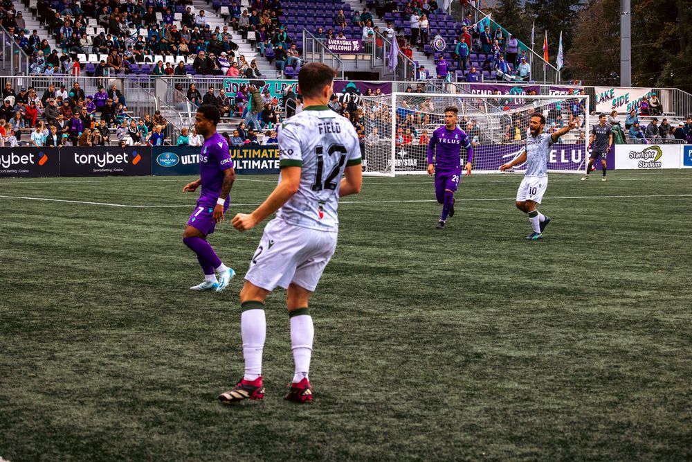 Soccer players compete on a grass field during a match with spectators watching from the stands in the background.