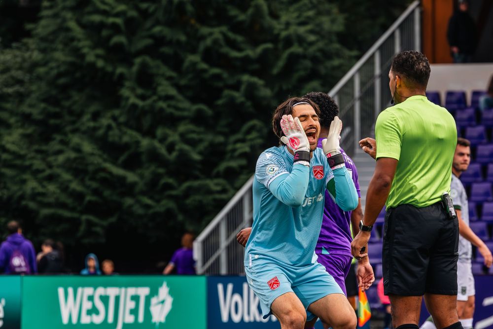 Soccer goalkeeper in light blue uniform reacts during match against team in purple on outdoor field.