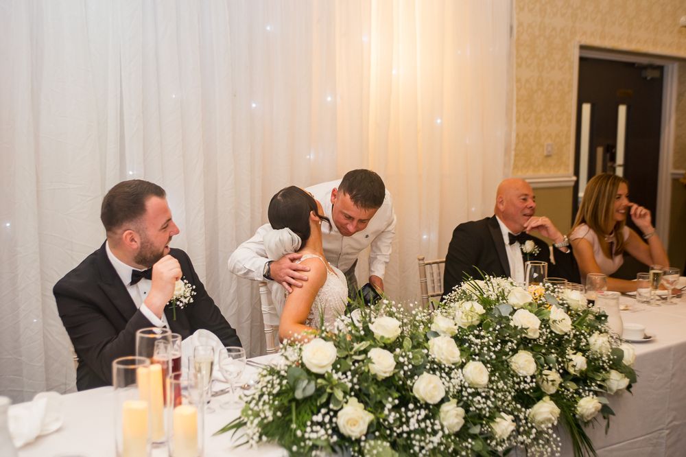 Wedding reception guests share laughter at an elegant head table decorated with white floral arrangements.