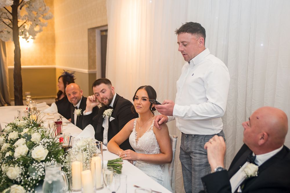 Wedding party enjoys entertaining speeches at reception with white floral centerpieces and candlelight.