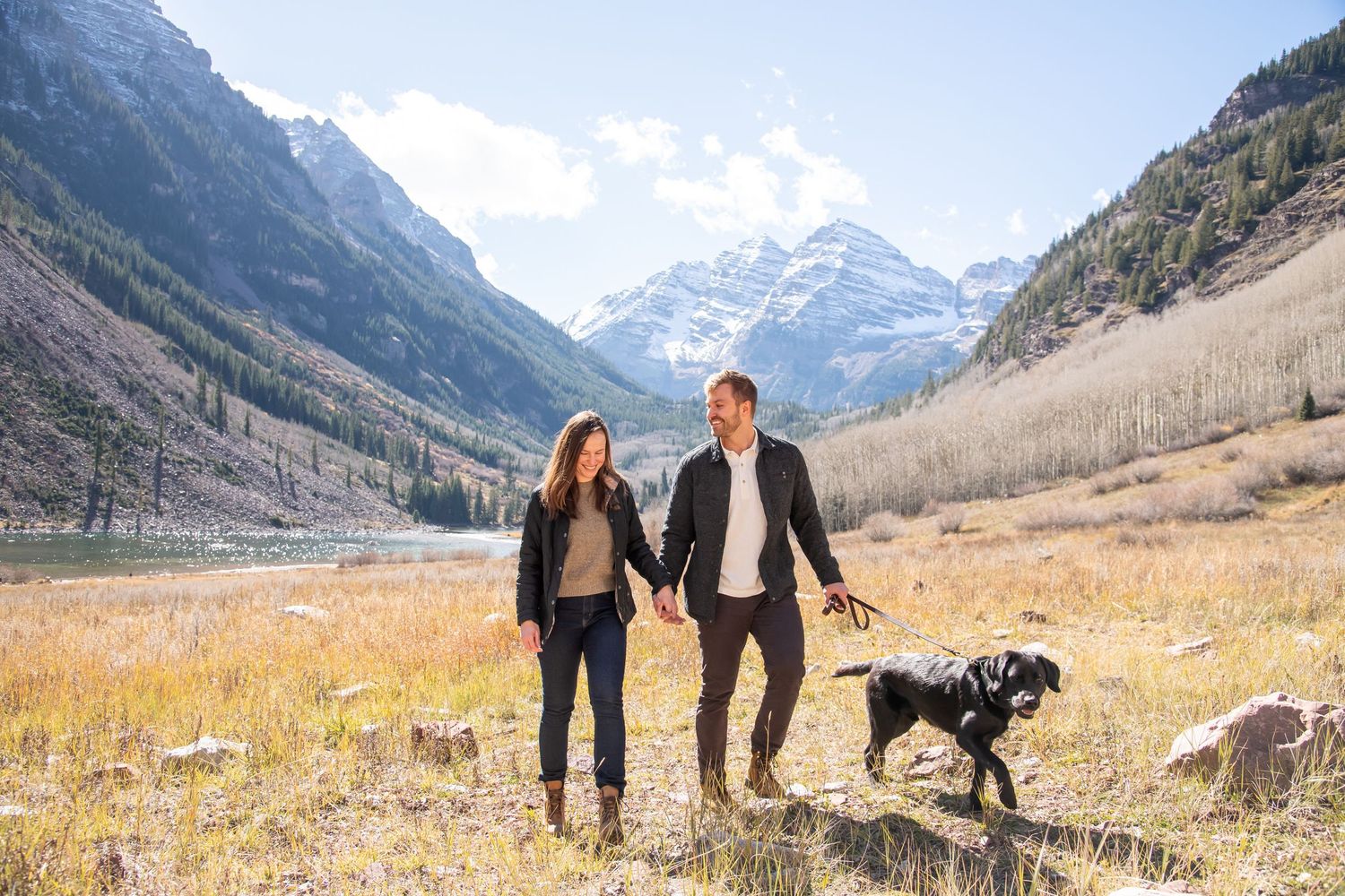 Cute candid image of a couple walking through a field with their dog at The Maroon Bells in Aspen Colorado, just after a surprise proposal
