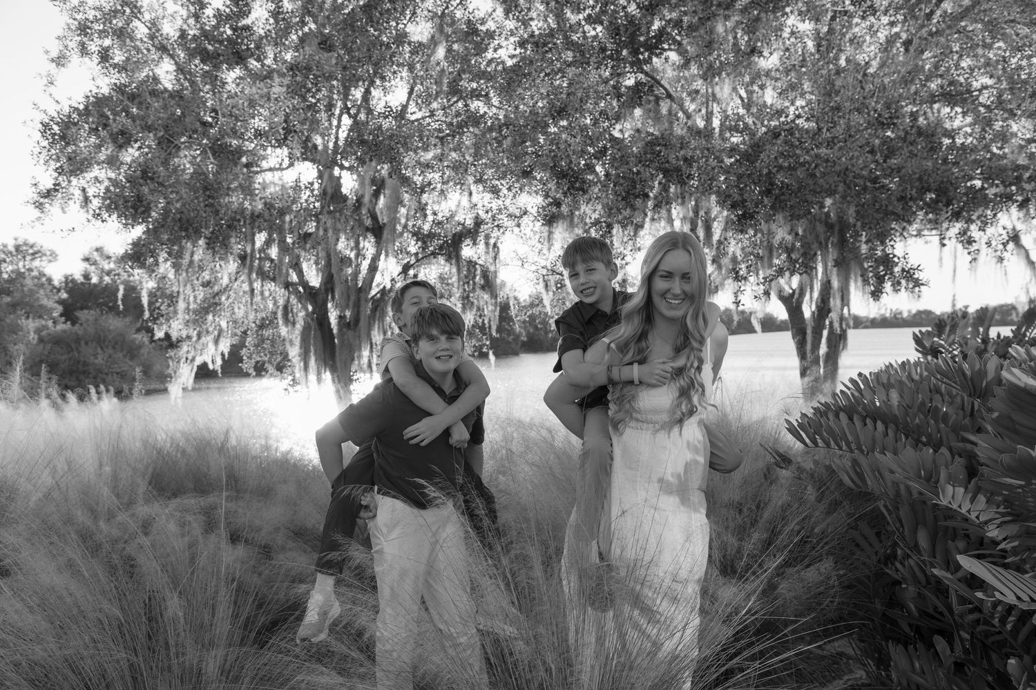 A woman in a white dress laughs as two boys playfully climb on her back near tall grass and mossy trees.