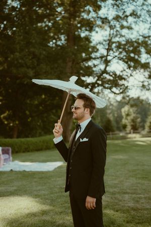 A person in a black suit holds a white parasol while standing on a lawn during an outdoor event.