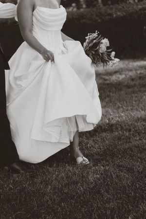 Black and white photo of a flowing white wedding dress and bouquet as someone walks on grass.