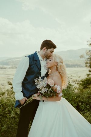 A romantic wedding couple embraces on a hilltop overlooking a scenic mountain landscape at sunset.