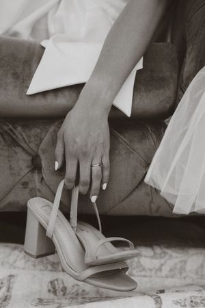 Black and white photo of a hand wearing a wedding ring reaching for an elegant heeled sandal on a velvet couch.