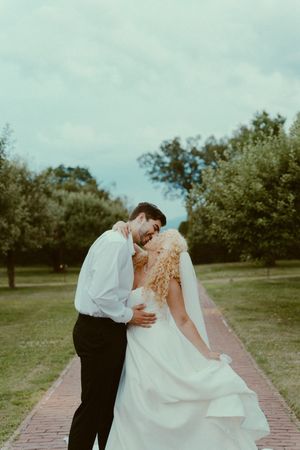 A romantic wedding couple shares a kiss while dipping on a brick pathway surrounded by green trees.