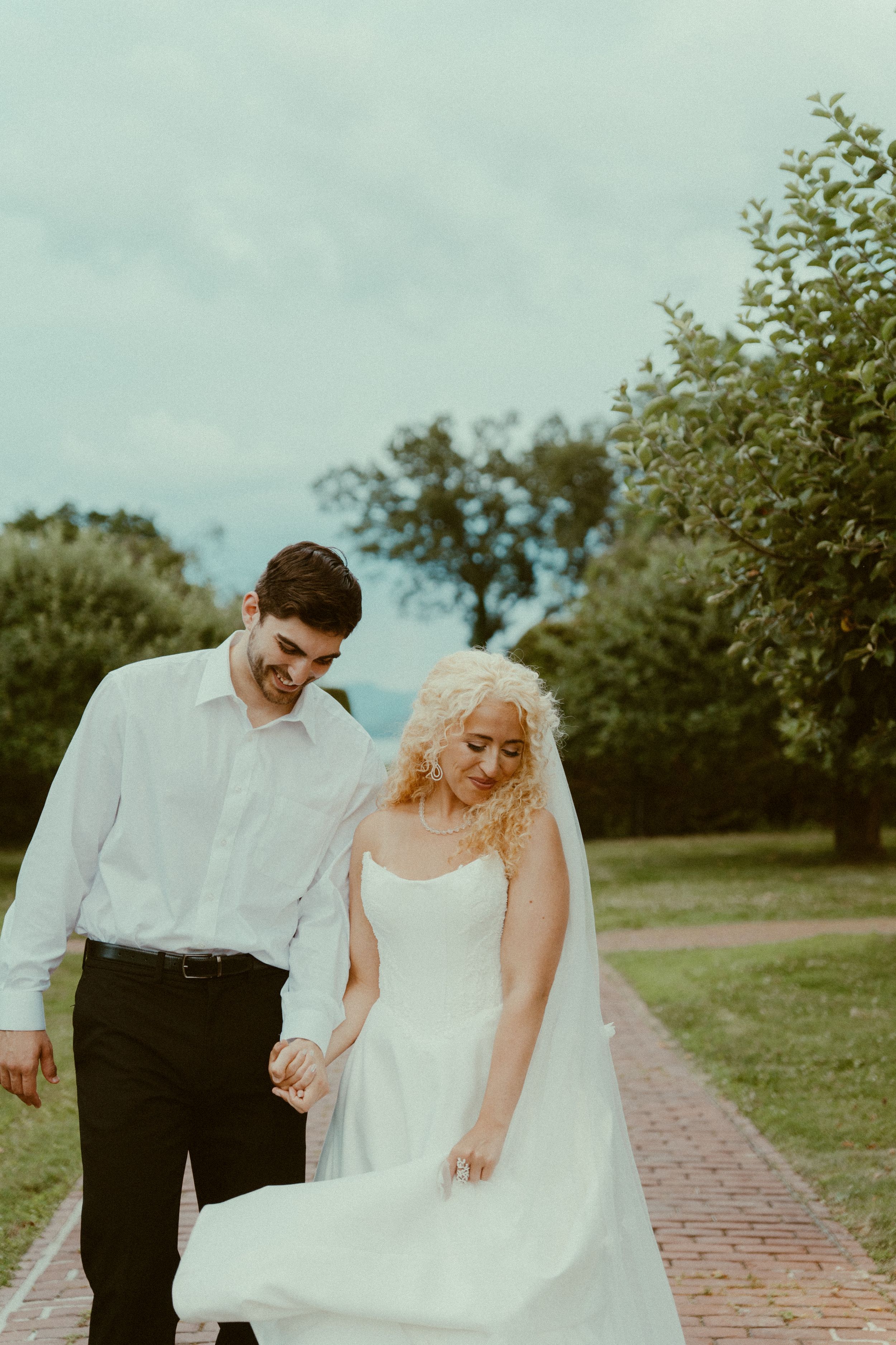 A couple in wedding attire walks together on a brick path surrounded by green trees and grass.