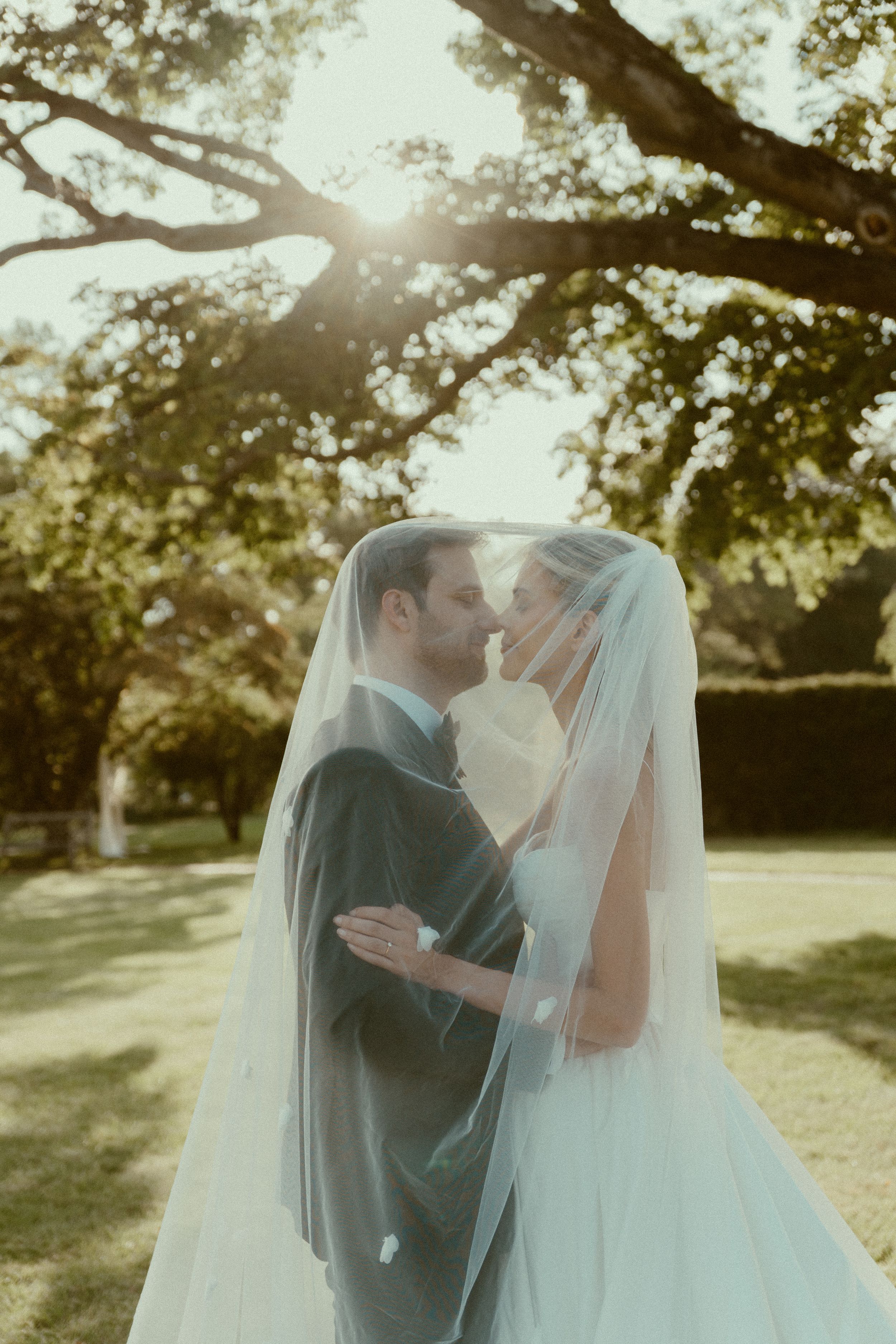 A romantic wedding couple shares an intimate kiss under sunlit trees during their outdoor ceremony.