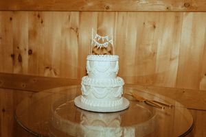 A three-tiered white wedding cake with elegant piping decorations sits on a glass stand against a wooden backdrop.
