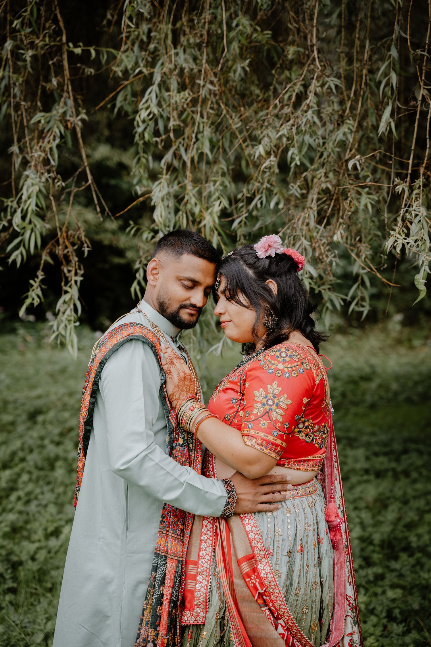 A romantic couple in traditional Indian wedding attire embrace under willow trees in an outdoor garden setting.