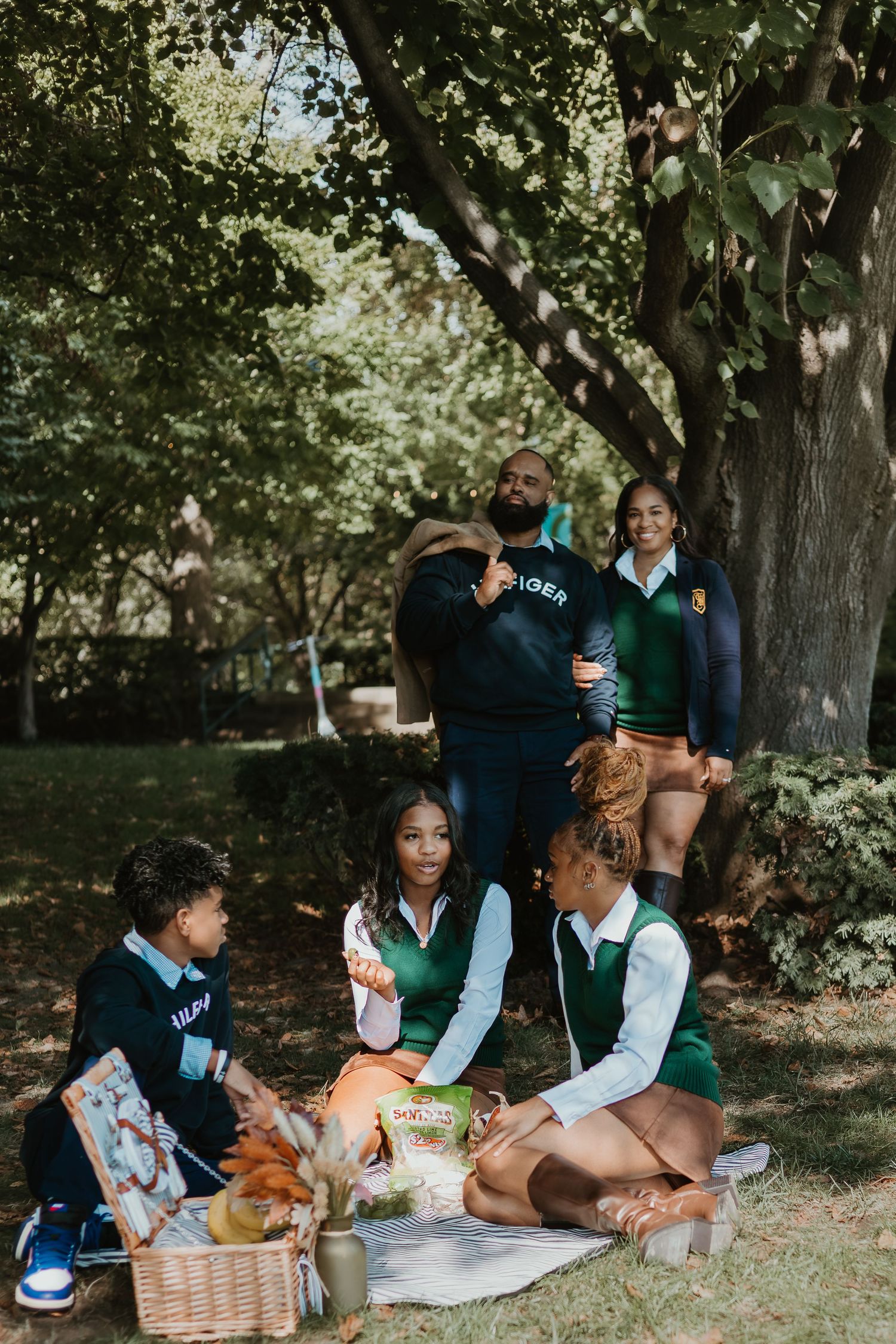 A group enjoys a sunny afternoon picnic together under shady trees in a park setting.