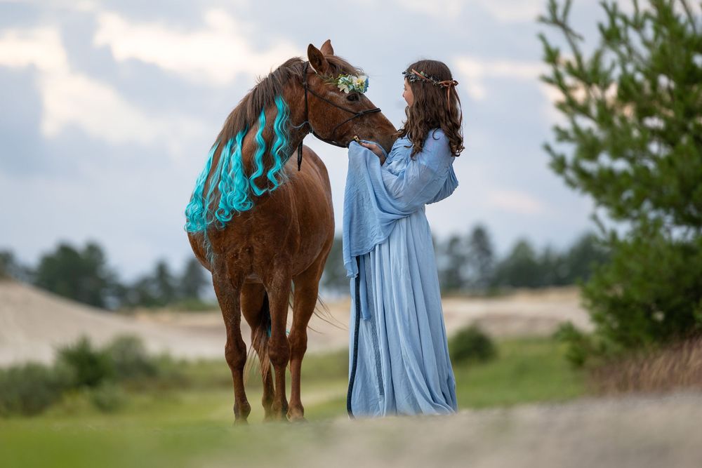 A person in a flowing blue dress dances in a field alongside a brown horse on a sunny day.