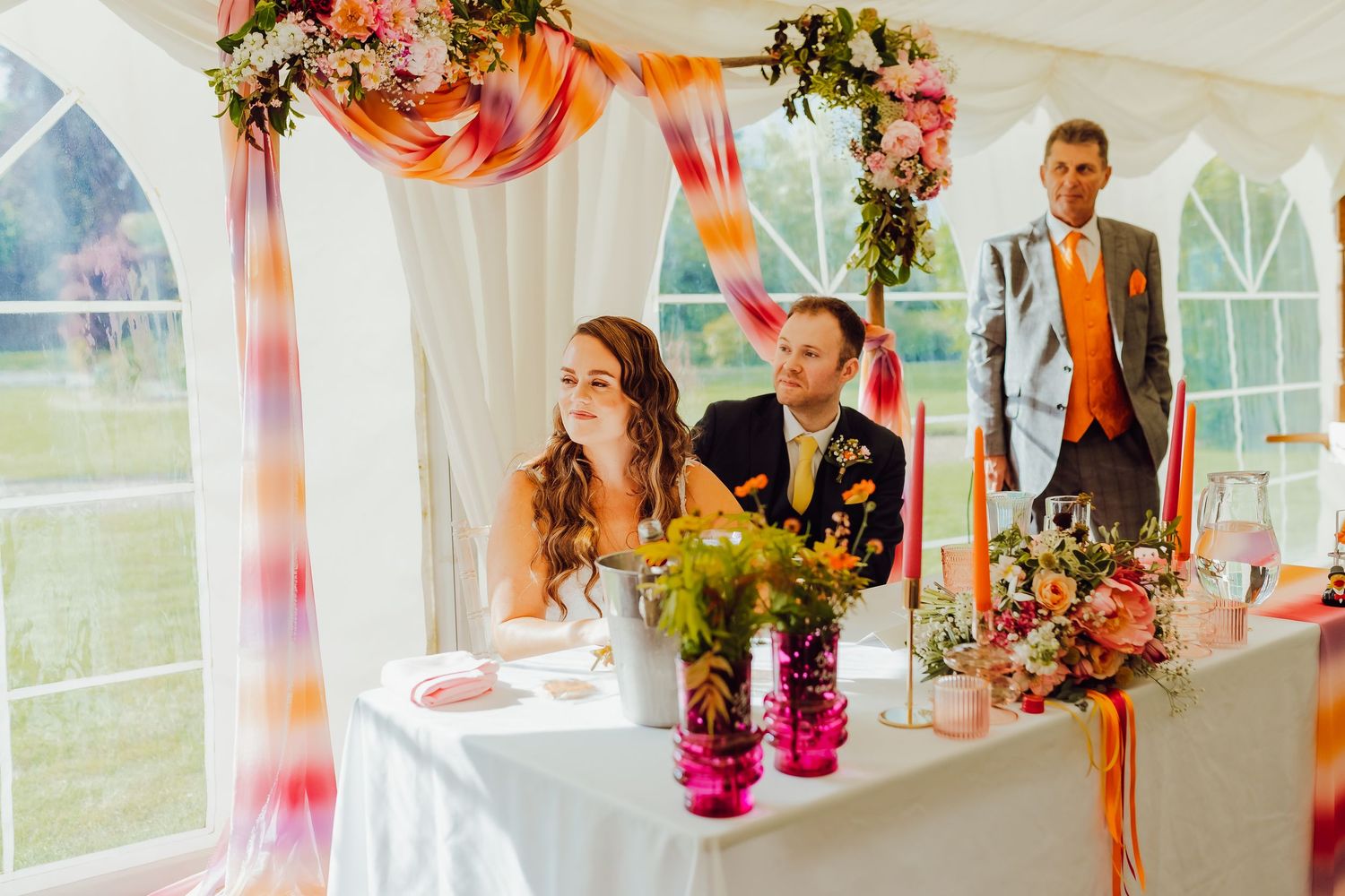 The wedding couple listening and smiling during speeches at their reception, enjoying the moment.