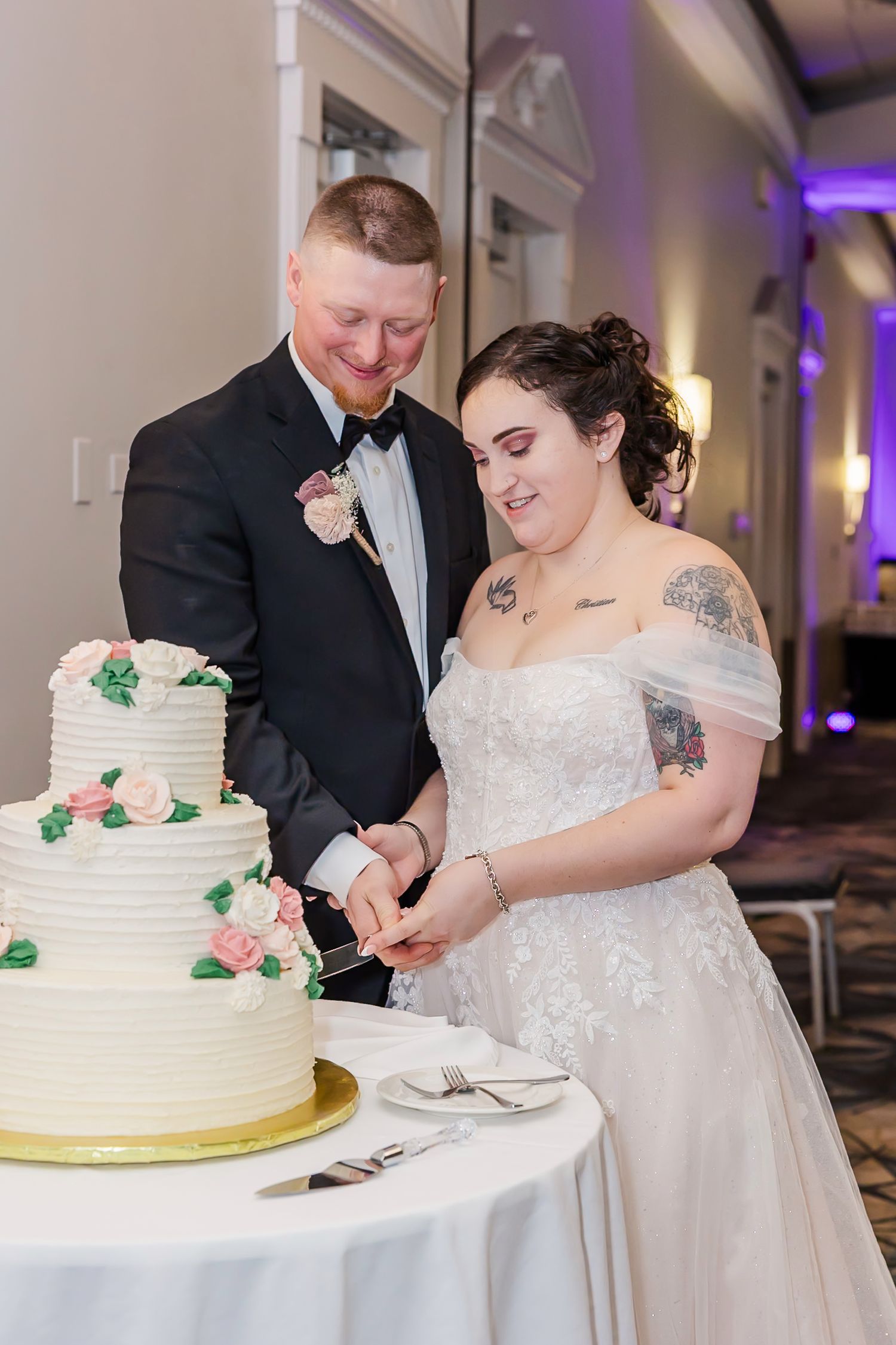 A couple in formal wedding attire cuts into their white tiered wedding cake decorated with green and pink accents.