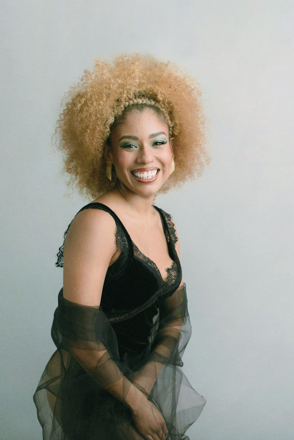 A figure in a black dress with sheer sleeves and voluminous blonde curly hair poses against a light backdrop.