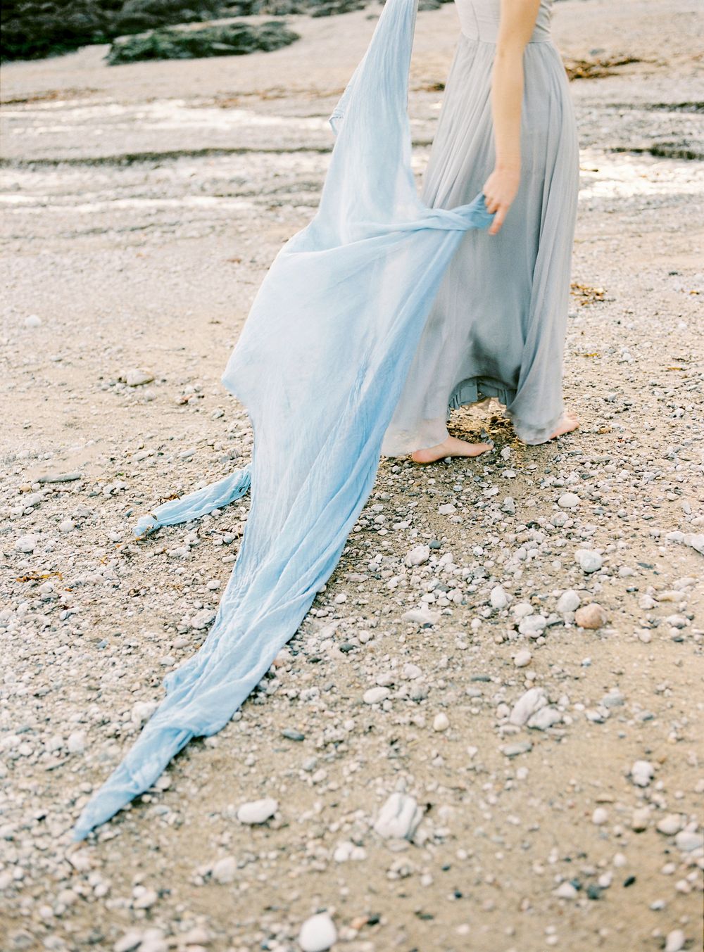 Flowing light blue chiffon dress train trailing on rocky ground creates ethereal movement in outdoor setting.